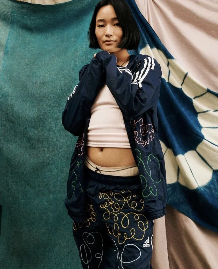 A young woman with shoulder-length black hair wearing a black and white Adidas tracksuit and beige crop top standing in front of colorful fabric drapes. The tracksuit is chainstich embroidered with swirly lines.
