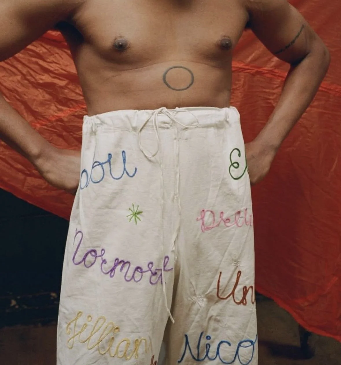 A shirtless man with tattooed arms and a circle tattoo on his abdomen, wearing light-colored shorts with handwritten colorful words and names, standing with hands on hips against a red backdrop.