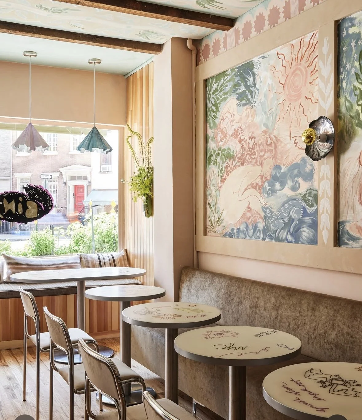 Interior of a cozy café with small round tables, upholstered seating, and a large window with a view of a city street. Decor includes wall art, hanging lamps, and a vase with flowers.