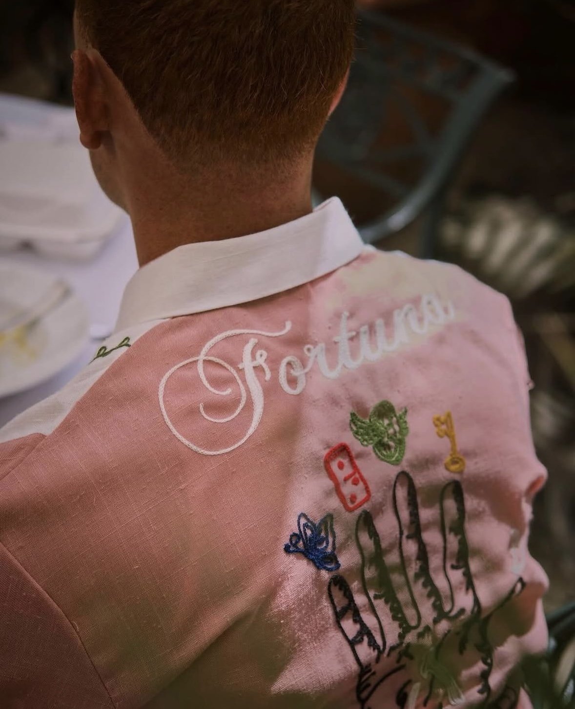 Back of a person's pink shirt with embroidered words 'Fortune' and various colorful embroidered illustrations including a green face, a red face, a yellow key, and other symbols.