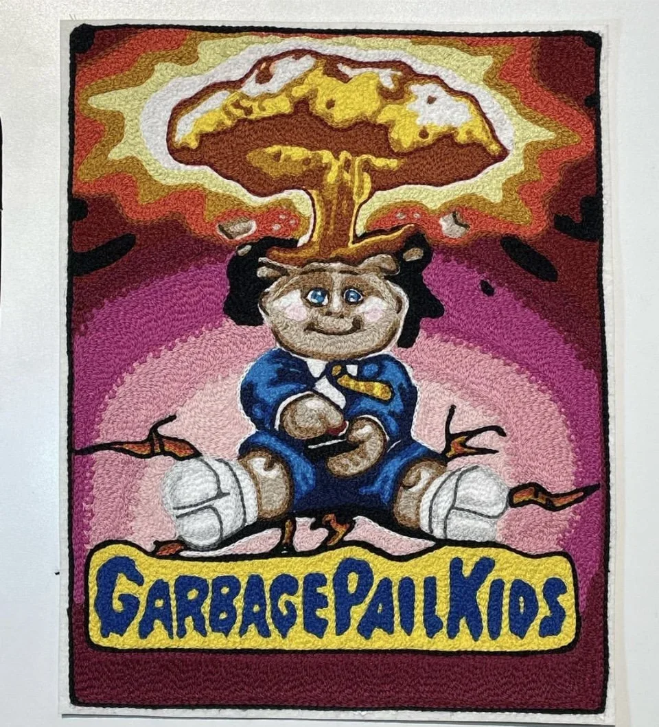 A childlike cartoon of a kid with blue eyes, wearing a blue jacket, sitting cross-legged, with a mischievous smile, manifesting a mushroom cloud explosion from their head. The background is pink and purple, with the words "GARBAGE PAIL KIDS" in yello