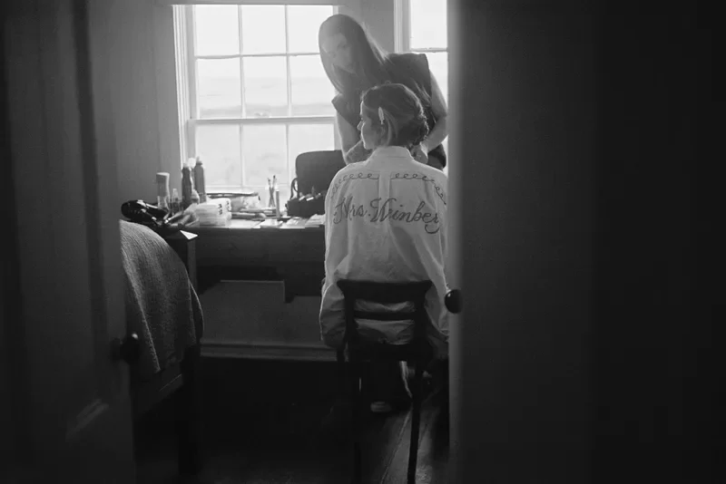 A bride sitting in a chair getting her hair done by a stylist in a room with a window, while a person stands nearby, both looking at her.