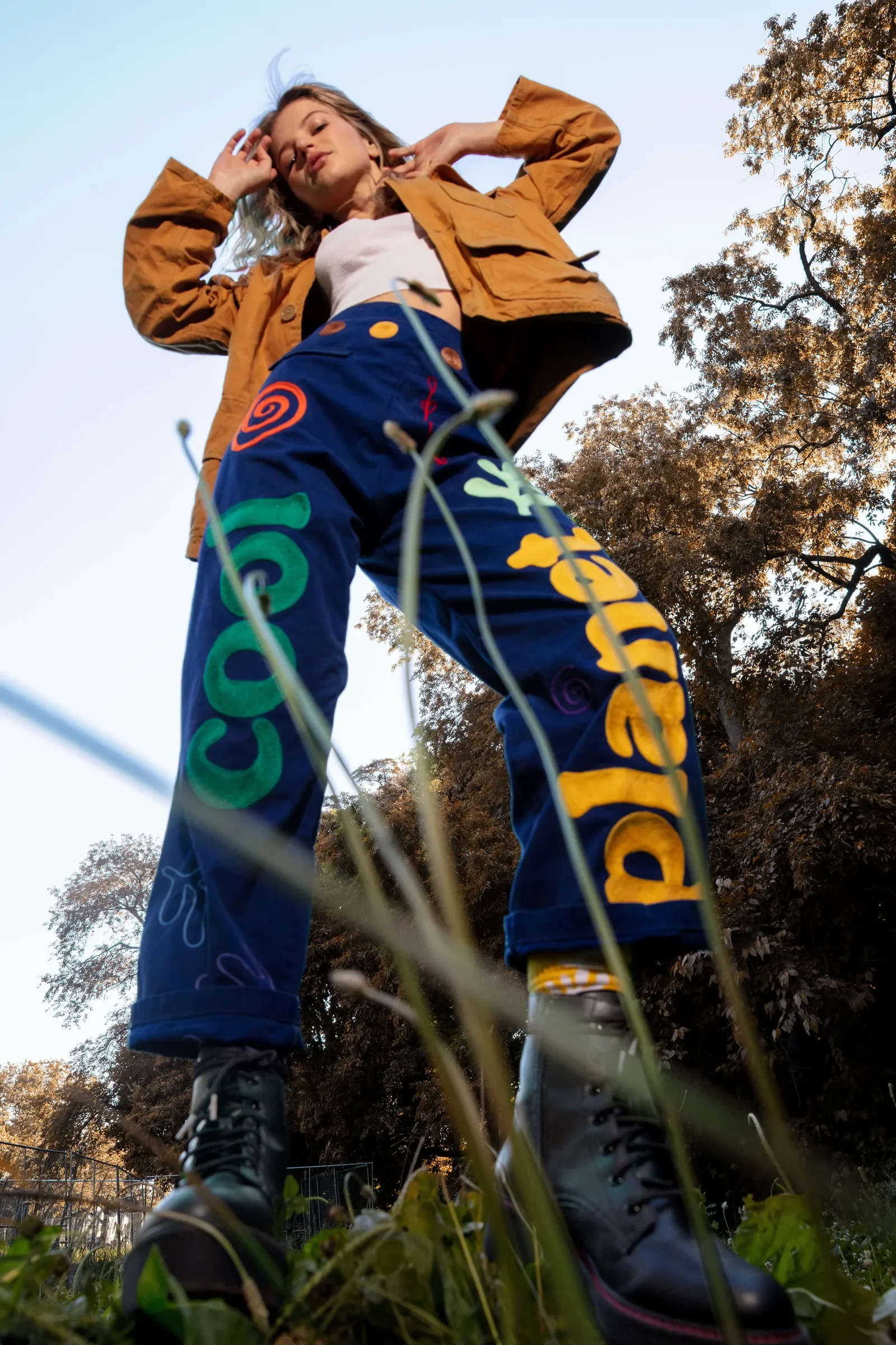 Low-angle shot of a woman standing outdoors, wearing colorful pants with the word 'Courage' on one leg and 'Dream' on the other, a white crop top, an orange jacket, and white shoes, with trees and a clear sky in the background.