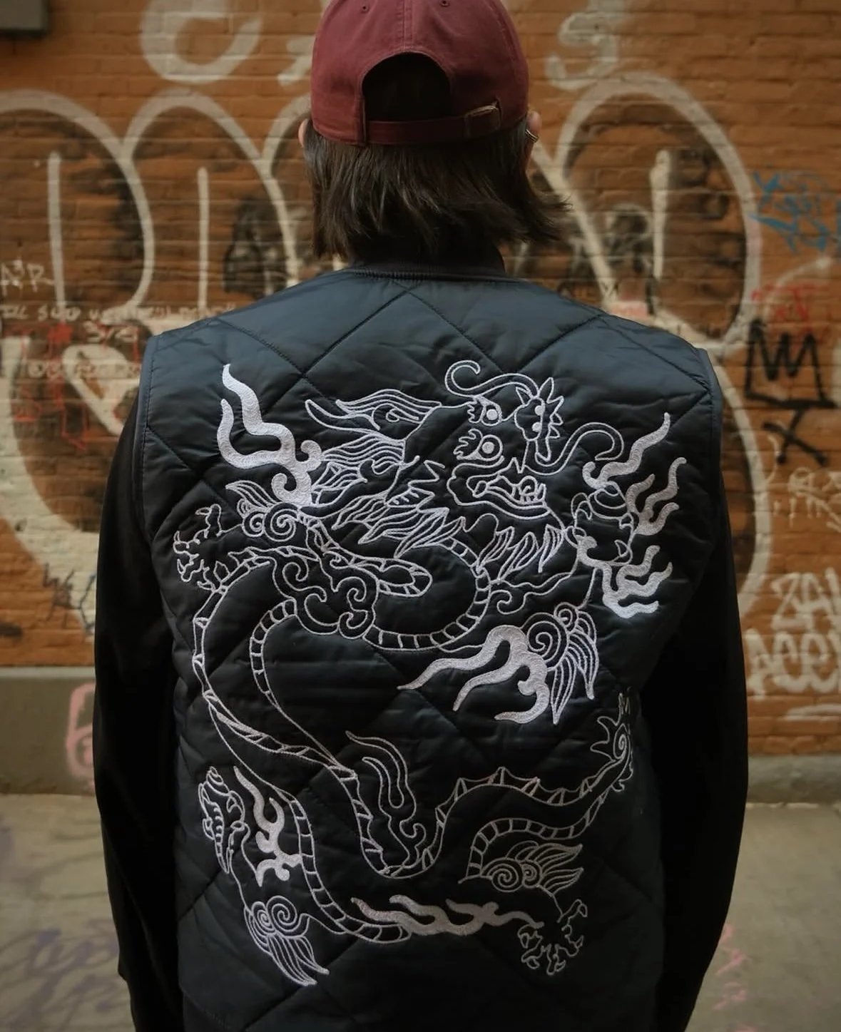 Person with long dark hair wearing a dark beanie and a black jacket with a white embroidered dragon design on the back, standing in front of a brick wall with graffiti.
