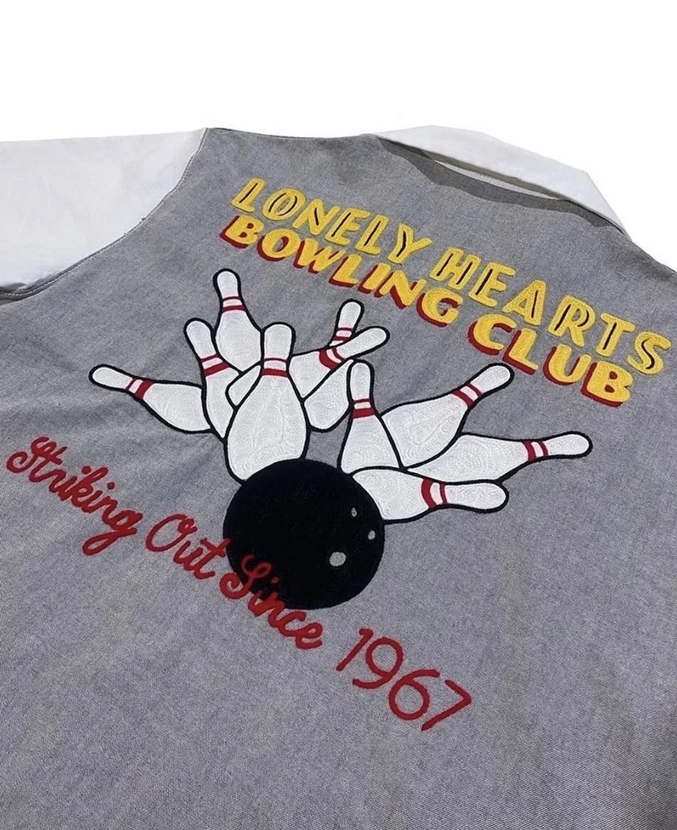 Gray bowling club shirt with a graphic of a bowling ball hitting bowling pins, yellow and red text that says 'Lovely Hearts Bowling Club', and red cursive text saying 'Striking Out Since 1967'.