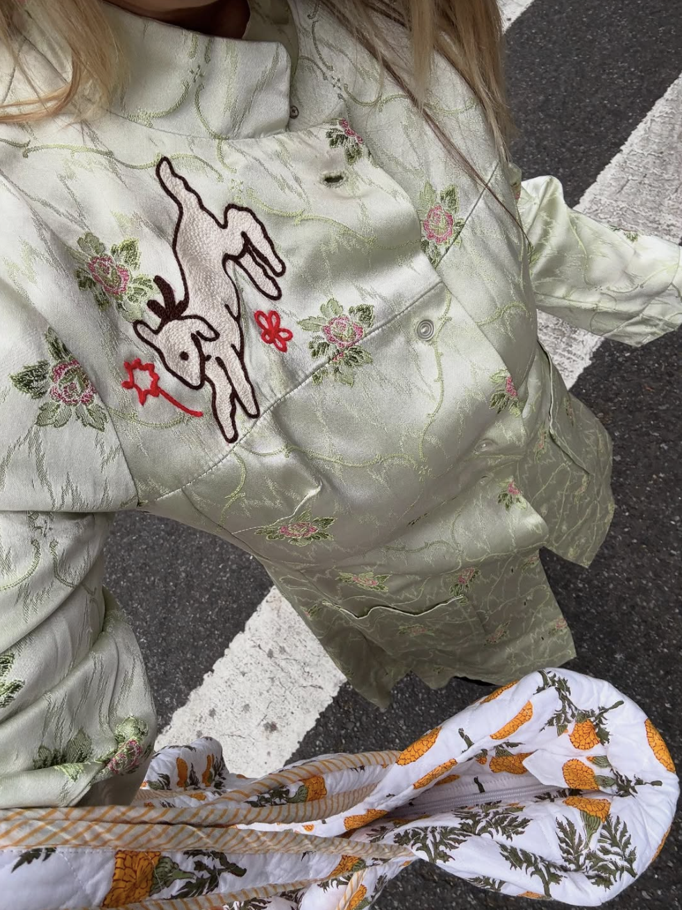 Person wearing a satin floral embroidered shirt with a donkey and flowers on it, standing on a street with a painted crosswalk.