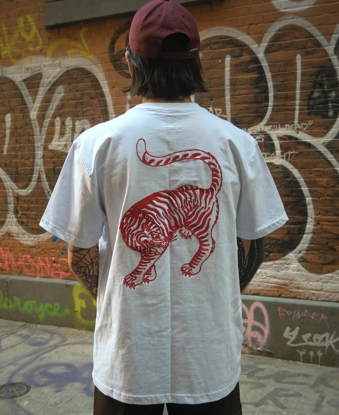 Person with shoulder-length dark hair wearing a maroon cap and white t-shirt with a large red tiger graphic on the back, standing in front of a graffiti-covered brick wall.