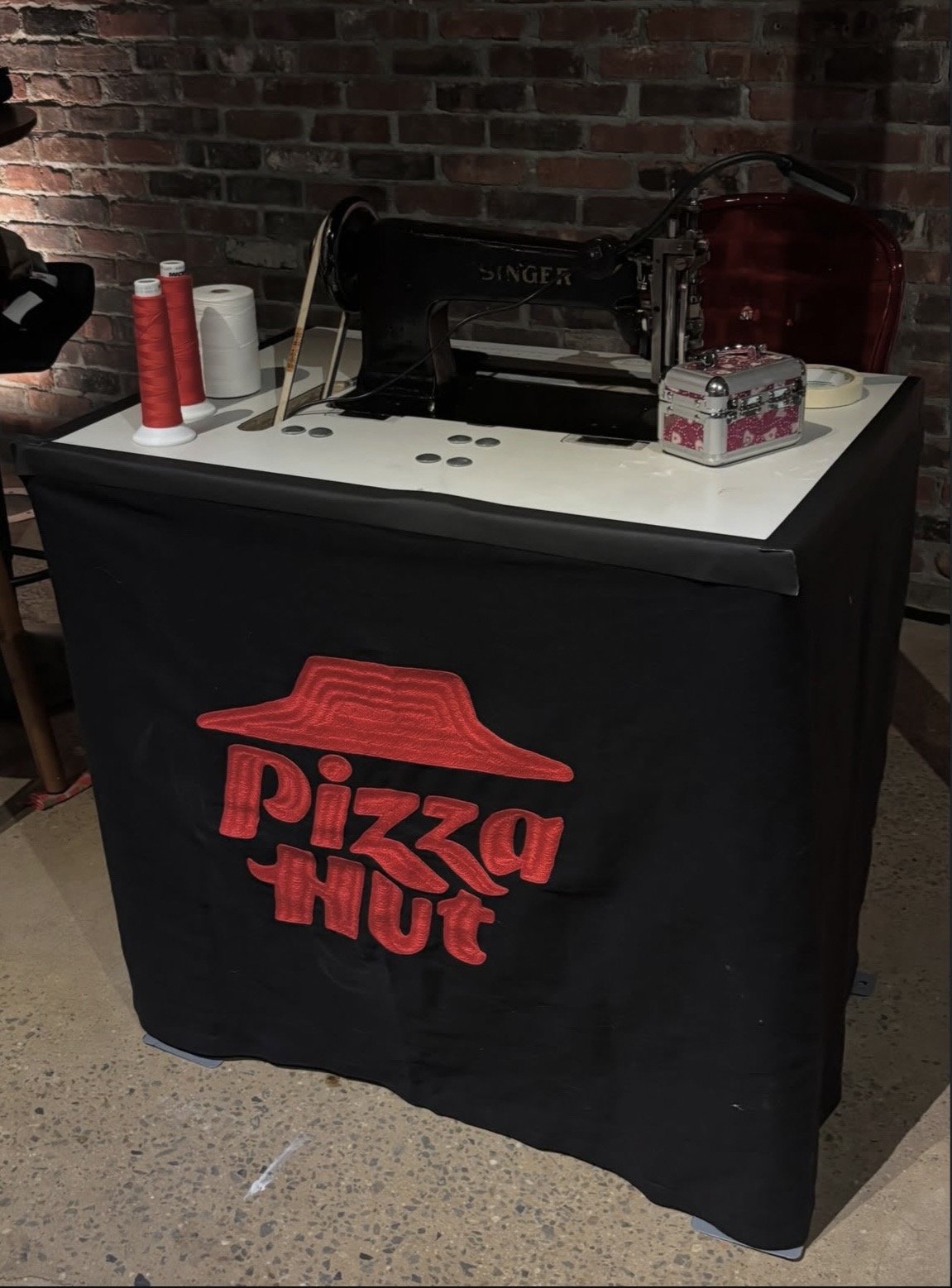A vintage Singer sewing machine on top of a black table with a pizza hut logo cover, decorated with sewing supplies including spools of red thread, fabric, and a small decorative box, against a brick wall background.