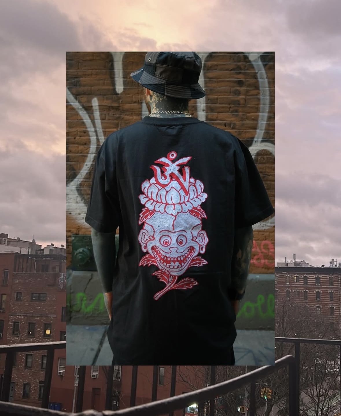 A person wearing a black bucket hat and a black shirt with a colorful clown face and flower design on the back, standing in front of a brick wall with graffiti, with an urban cityscape in the background during sunset.