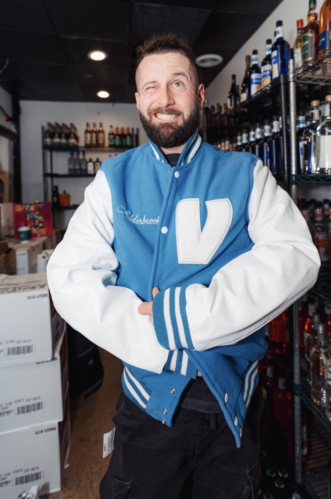 A man with a beard and short hair winking, wearing a blue and white varsity jacket with the name 'Elderbrook' and a large letter 'V' on it, standing in a liquor store with shelves of alcohol and boxes in the background.