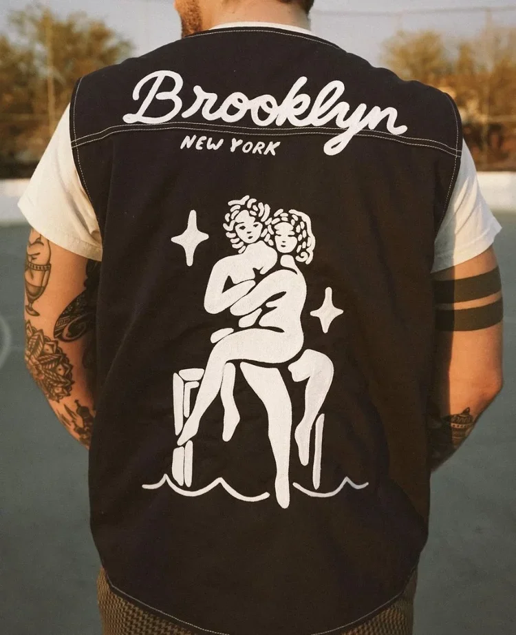 The back of a person wearing a black vest with white illustrations of two women sitting together, surrounded by stars and water. The vest has the words "Brooklyn New York" written at the top.