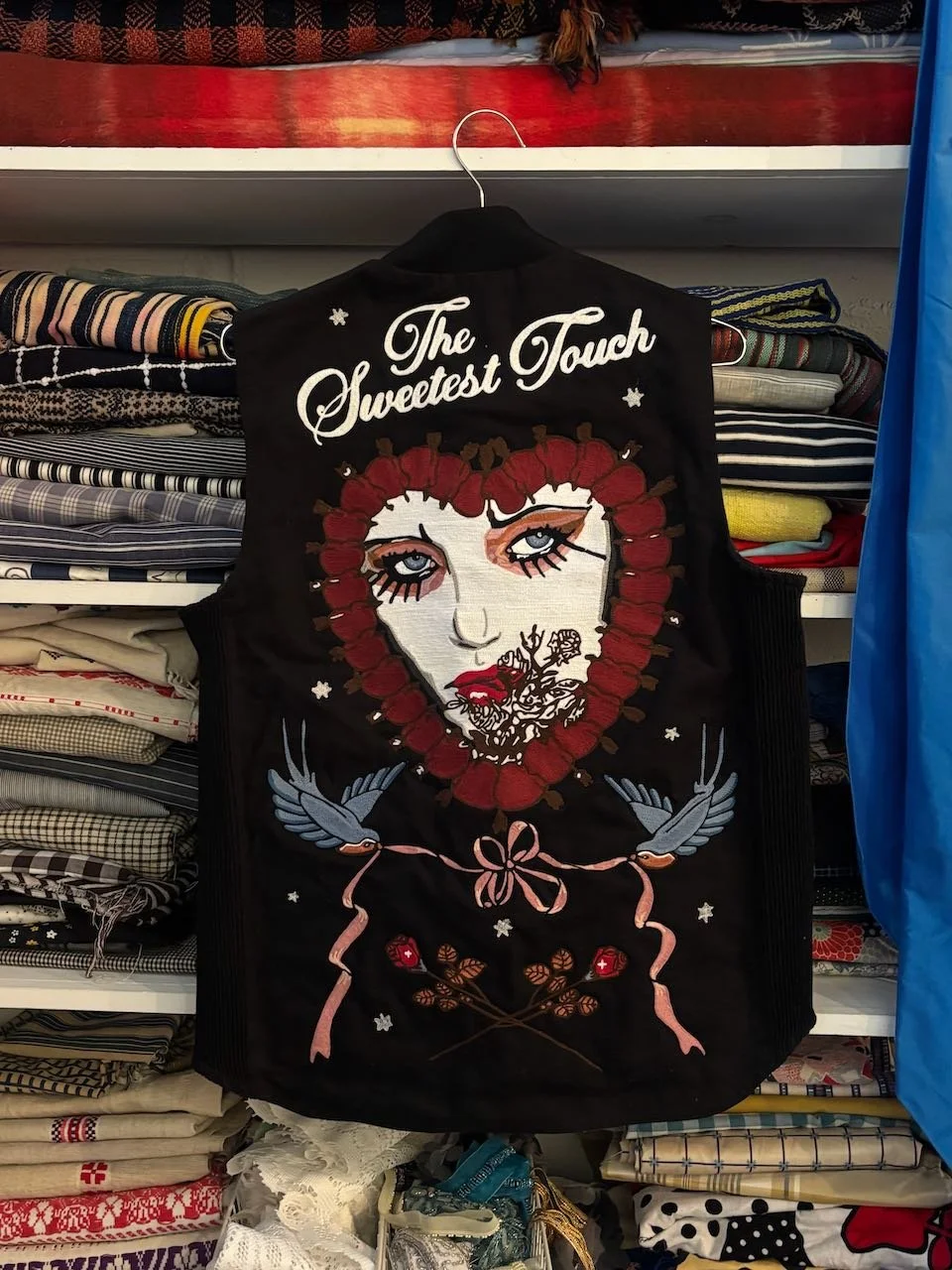 A black embroidered jacket with the words 'The Sweetest Touch' on the top. It features an embroidered face of a woman with red hair, blue eyes, and makeup, surrounded by floral and bird designs.