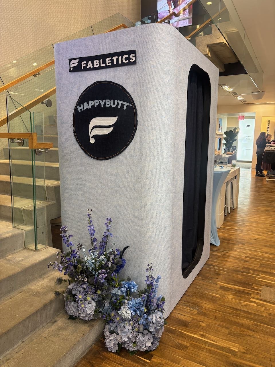 Indoor setup with a large fabric-covered photo booth, decorated with a sign that reads 'FABLETICS' and a circular logo with the text 'HAPPYBUTT' beside a stylized fabric swatch. There are purple and white flowers at the base of the booth beside a sta