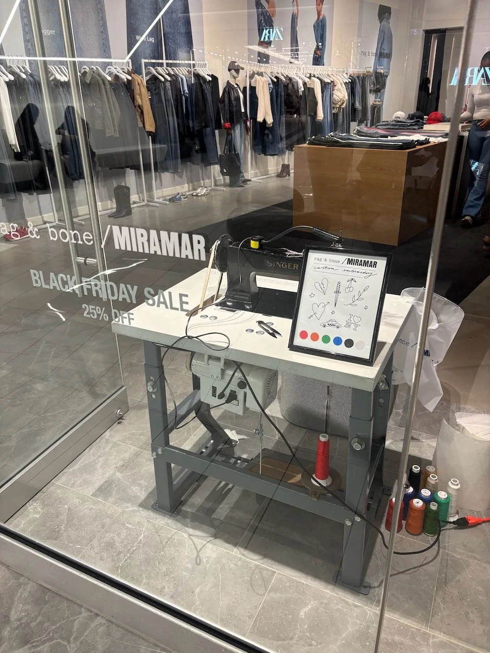 Empty checkout station at a clothing store, with a computer, scissors, and a framed drawing of hearts, umbrellas, and the words "I love". There are spools of thread on the floor, and clothing on racks behind the glass.