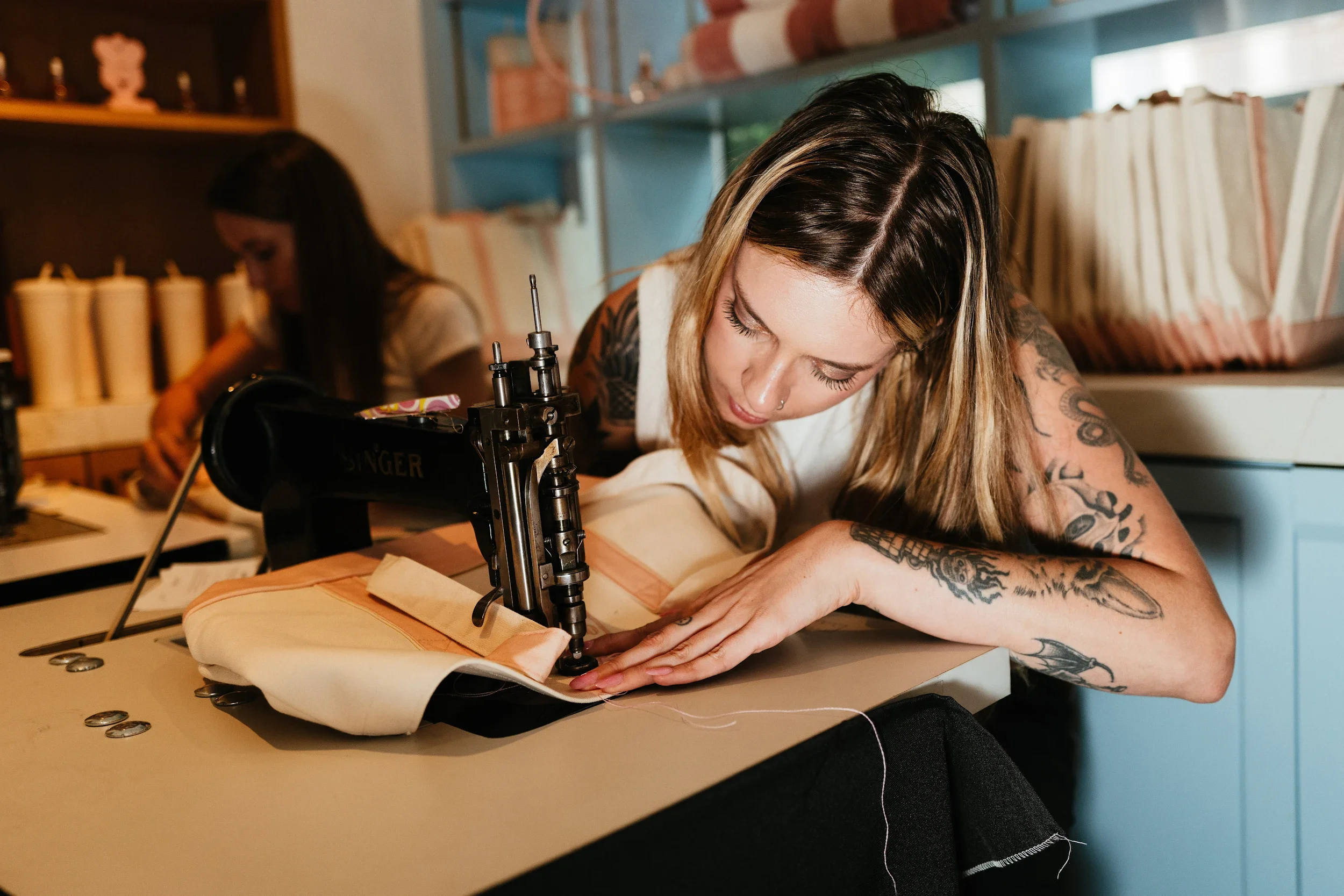 A woman with tattoos using a sewing machine to sew fabric on a worktable, with another woman in the background also working, in a room with shelves and books.