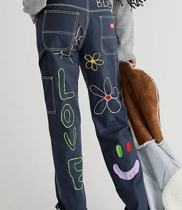 Close-up of a person wearing dark blue jeans with colorful embroidery, including flowers, the word "LOVE," a smiley face, and other decorative elements. The person is holding a brown item, possibly a stuffed toy or pillow, and is dressed in a gray knitted sweater.