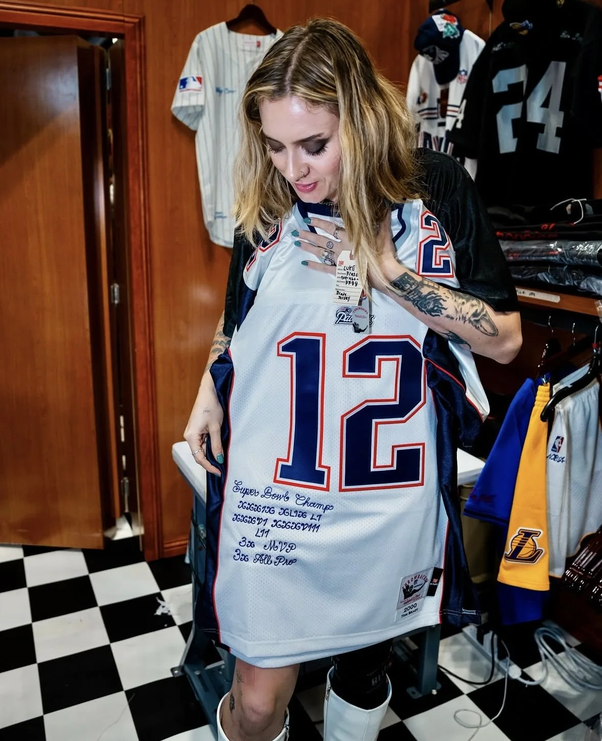 A woman with tattoos and blonde hair looking down at a New England Patriots jersey she is holding, in a room with sports jerseys hanging on the wall.