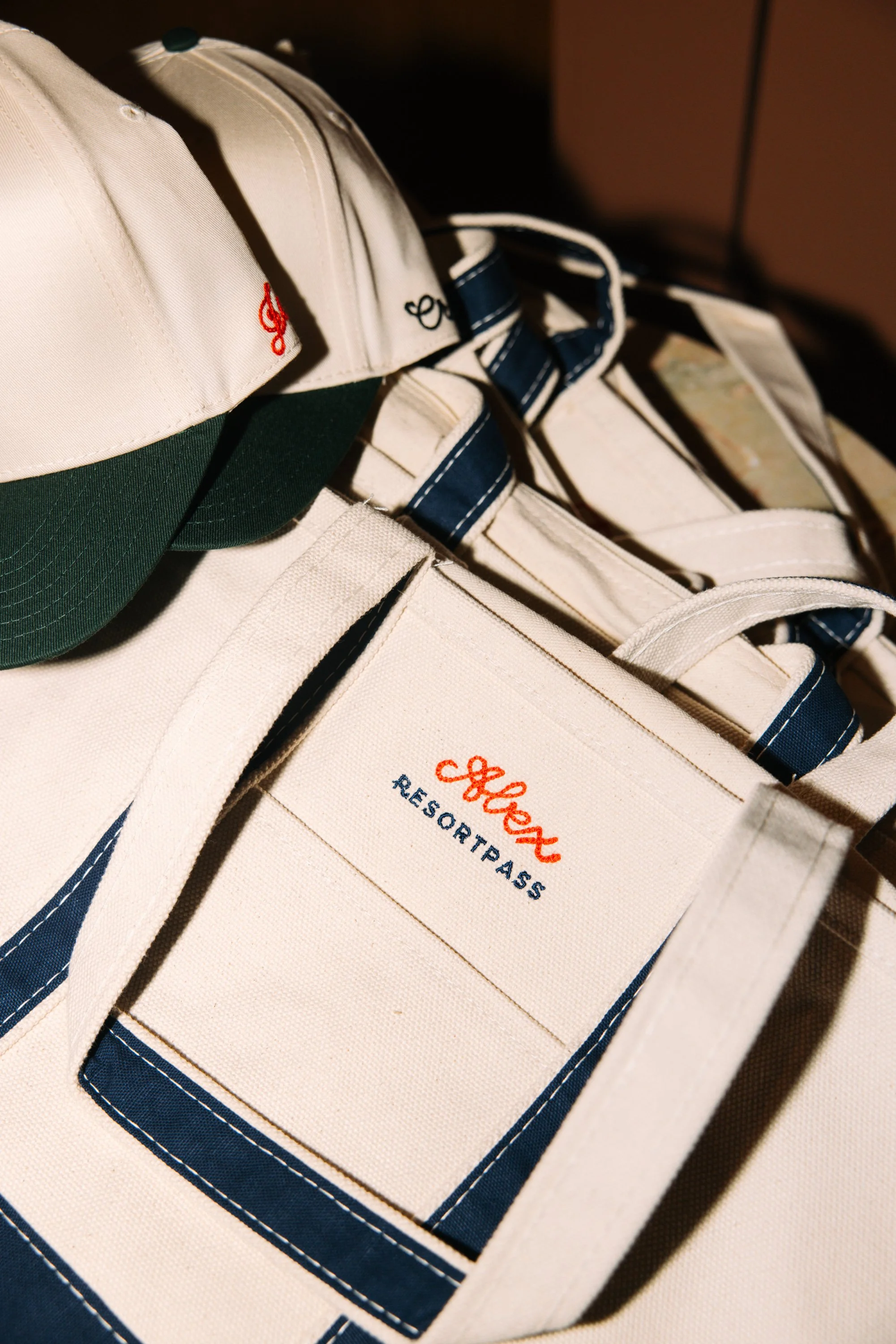 Close-up of white canvas Tote bag with navy accents, embroidered with orange and navy text, and a white baseball cap with a green brim and embroidered red and black text.