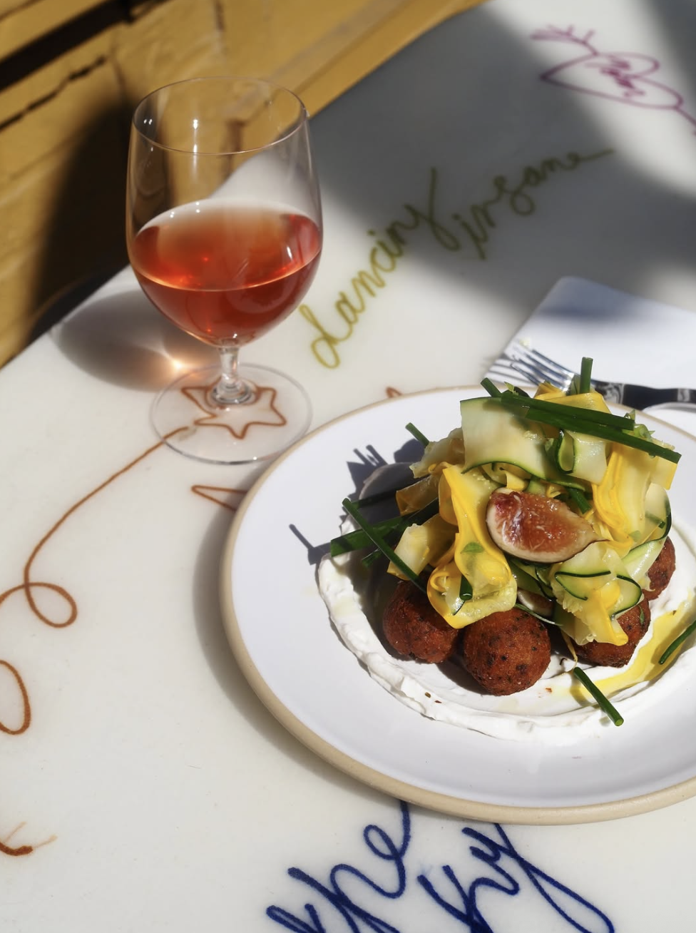 A glass of pinkish-red wine and a plate of food with falafel balls, garnished with sliced cucumbers, yellow bell peppers, chives, and a fig, on a white tablecloth with handwritten text.