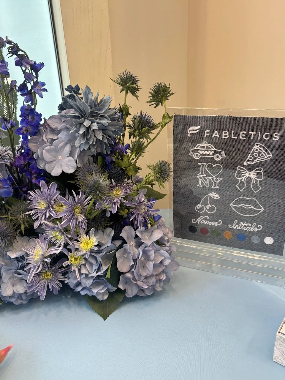 A floral arrangement with purple and blue flowers next to a black sign that reads "Fabletics" with line drawings of a car, pizza, hearts, cherries, lips, and the words "Names" and "Initials" on a light blue table near a window.