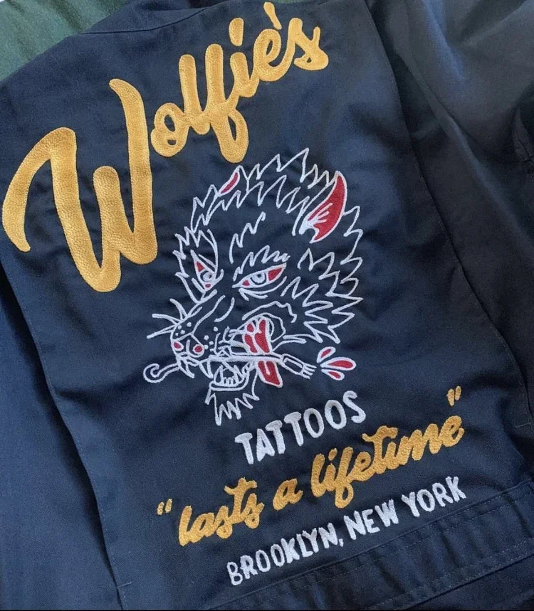 Backpack with embroidered logo of the tattoo shop 'Wolffie's Tattoos' in Brooklyn, New York, featuring a fierce wolf face holding a tattoo machine in its mouth, with the text 'lasts a lifetime' in yellow cursive.