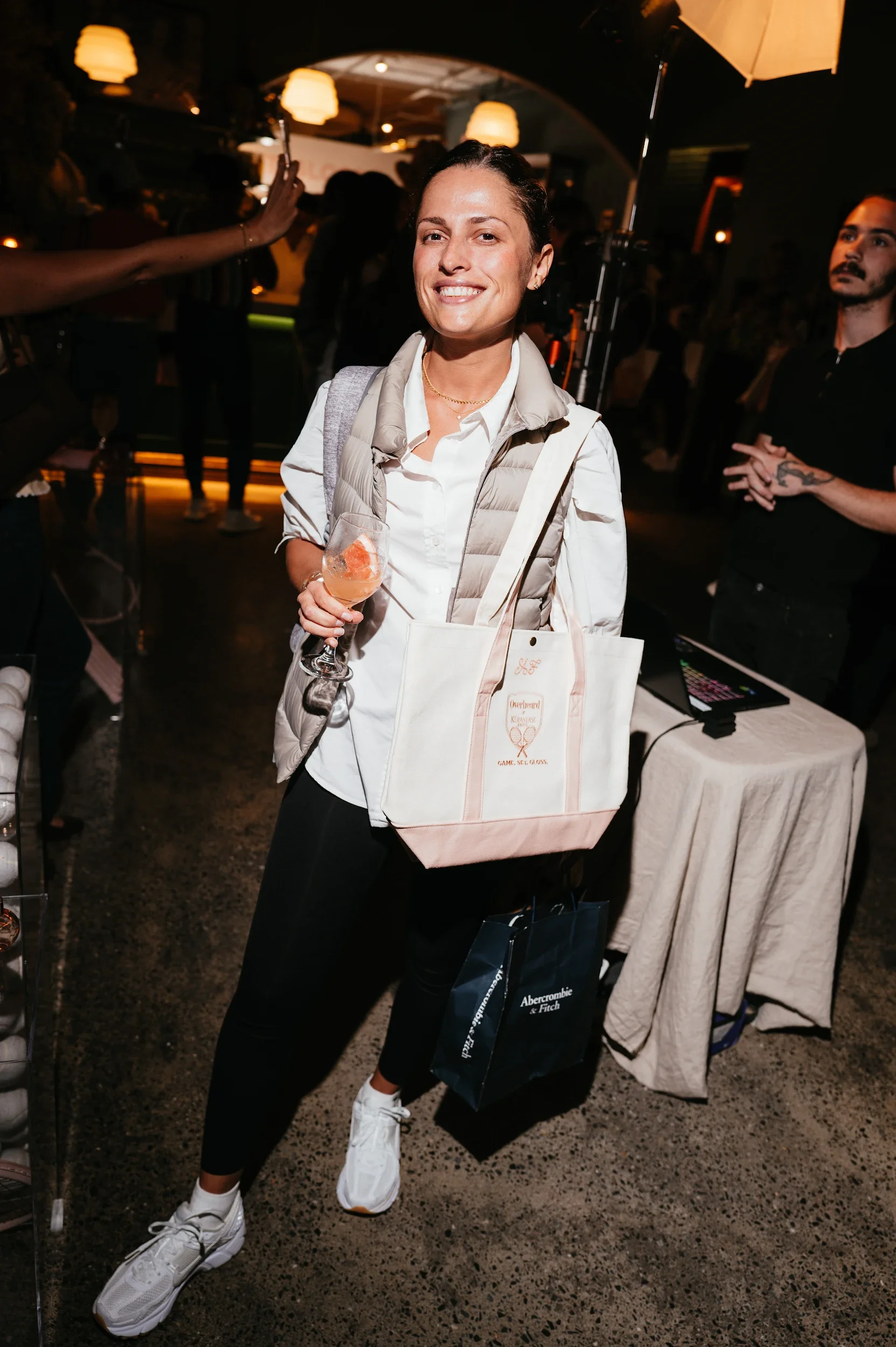 A woman smiling at a social event, holding a glass of drink, wearing a white shirt, black pants, and white sneakers. She has a beige vest and a tote bag with text, standing in a dimly lit room with other people and equipment around her.