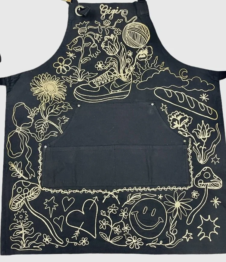 Black apron with yellow line drawings including flowers, a sneaker with a ball of yarn and crochet hook, bread, mushroom, a smiley face, hearts, stars, and abstract shapes.
