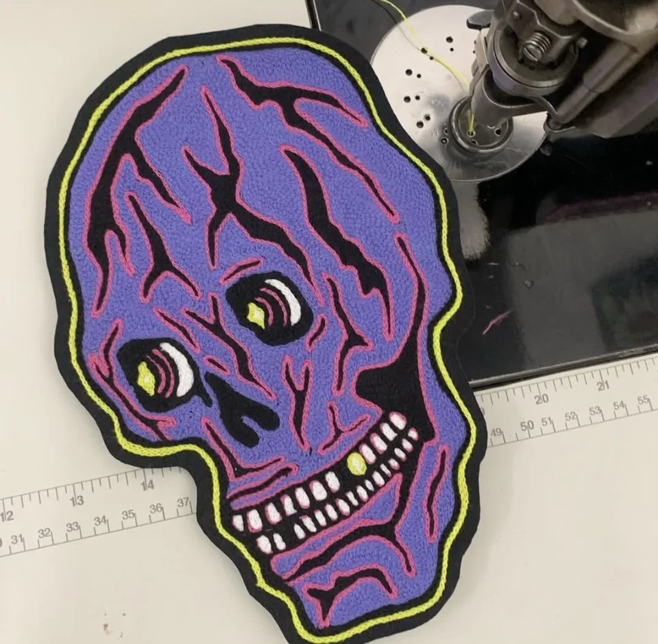 Colorful embroidered patch of a purple skull with black and pink outlines and yellow eyes, placed on a white surface with a ruler next to it.