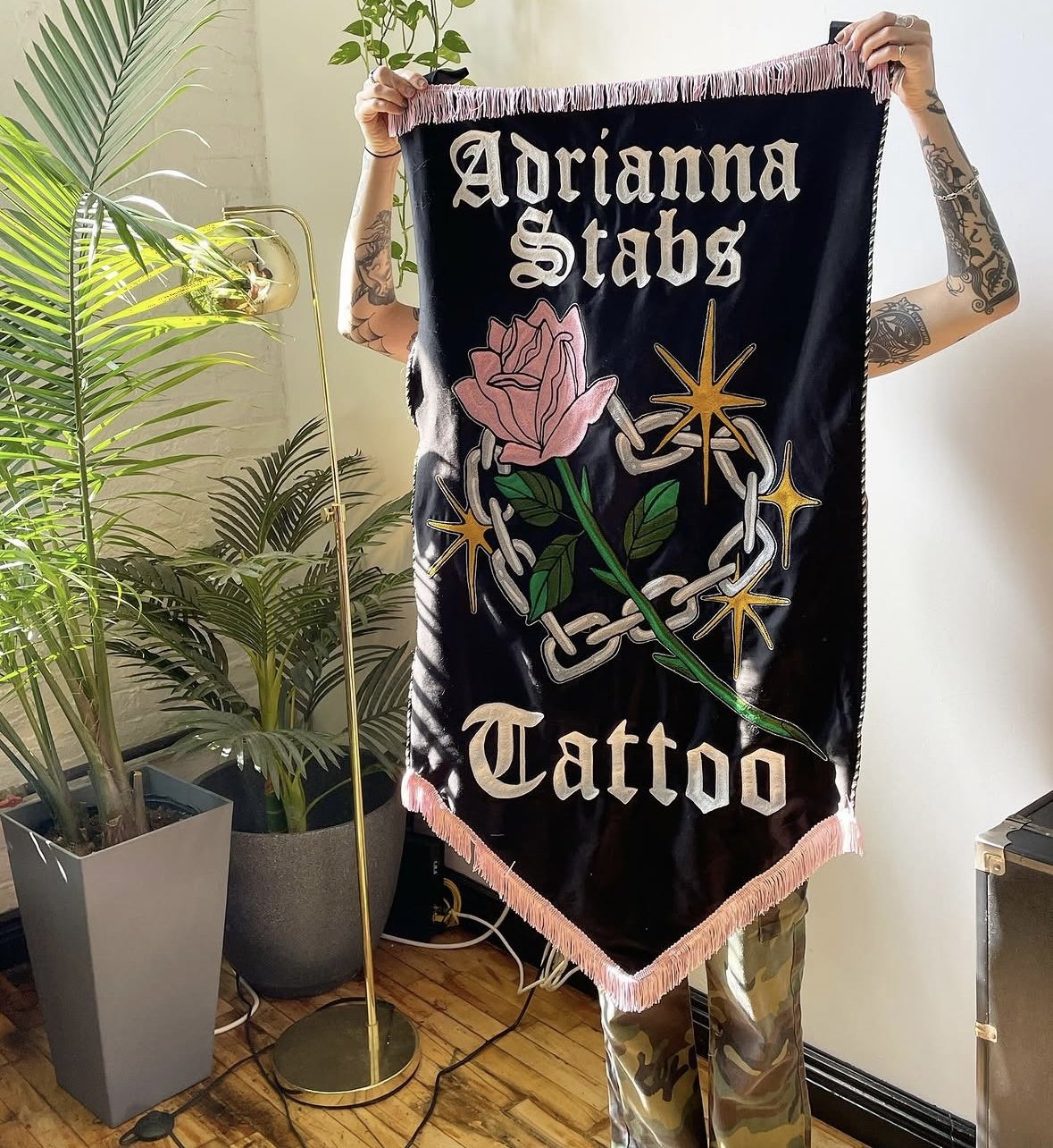 A person holding a chainstitched black banner that reads 'Adrianna Stabs Tattoo' with a pink rose, green leaves, and stylized stars and chains. The person has tattoos on their arms and is standing indoors with potted plants in the background.