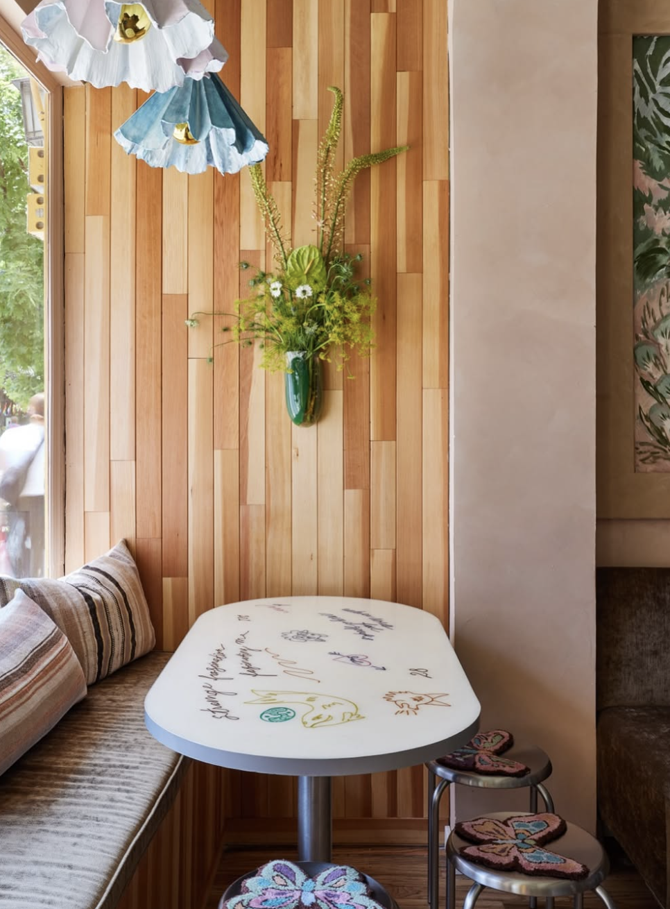 A cozy corner with a panel wooden wall, a green floral wall-mounted vase with a fresh flower arrangement, a white table with colorful chainstitch embroidery, a cushioned bench with pillows, and two round stools with butterfly-shaped cushions.