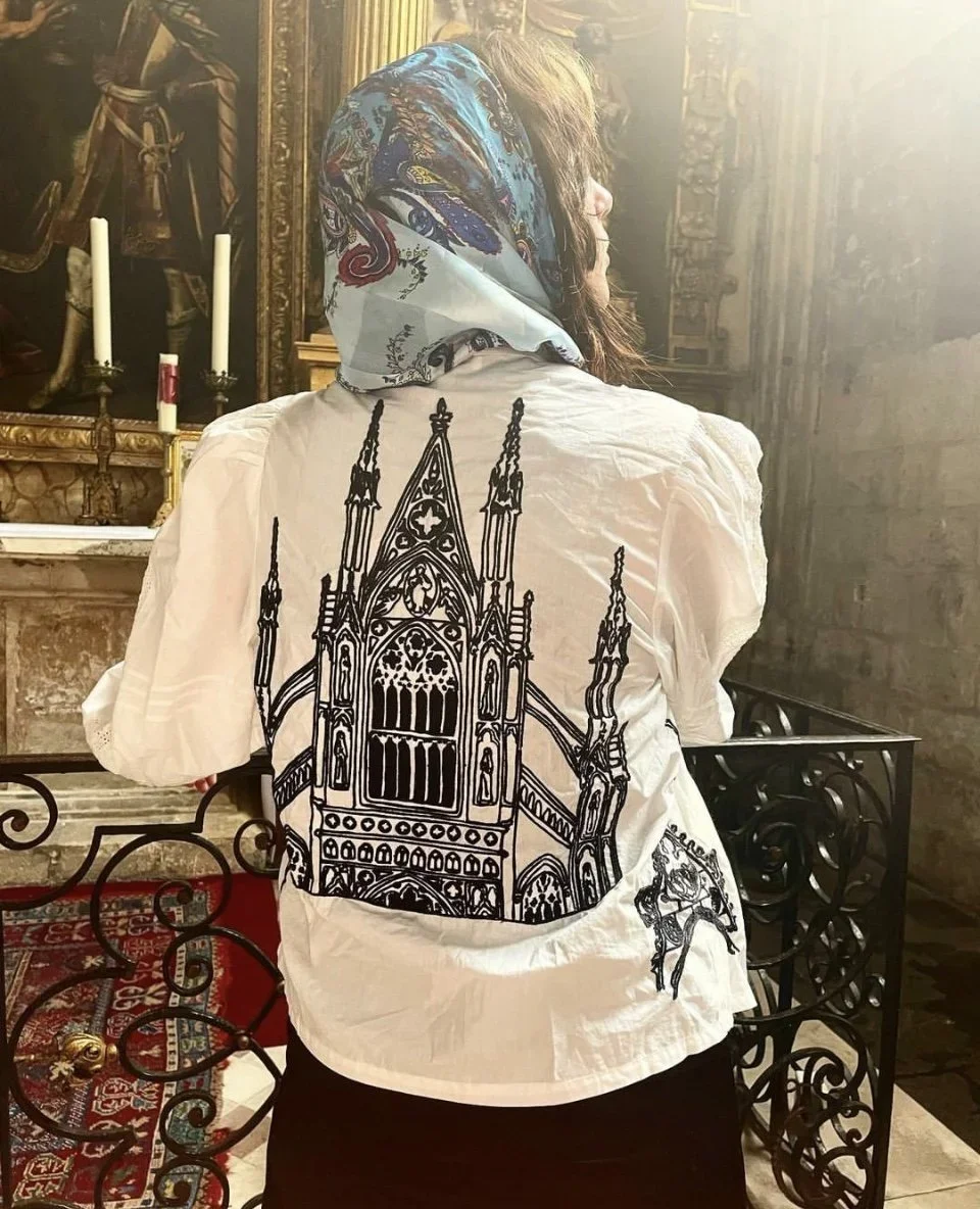 A woman with a colorful headscarf and white blouse with a black architectural design on the back, standing inside a church or historical building with ornate interior and religious artifacts.