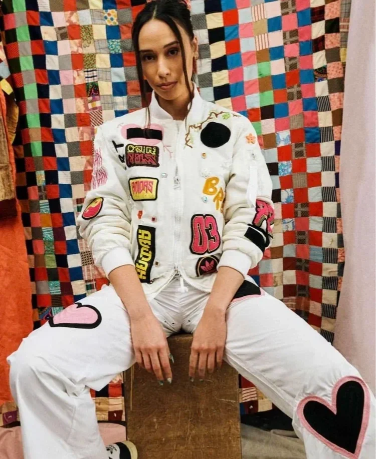 A young woman sitting on a wooden bench in front of a colorful patchwork quilt, wearing a white jacket and pants decorated with patches and chainstitched hearts.