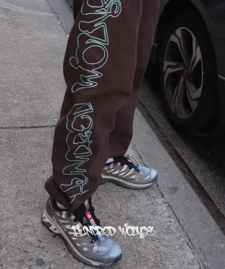 Person standing on a sidewalk near a parked car, wearing brown sweatpants with white text on the side and gray sneakers.