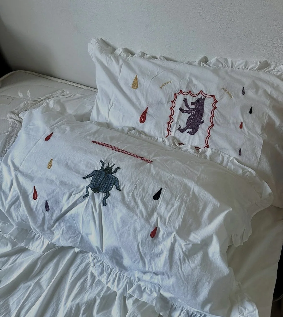 Decorative white bedding with embroidered bugs and raindrops, featuring a croc and a beetle, on a bed with ruffled edges.