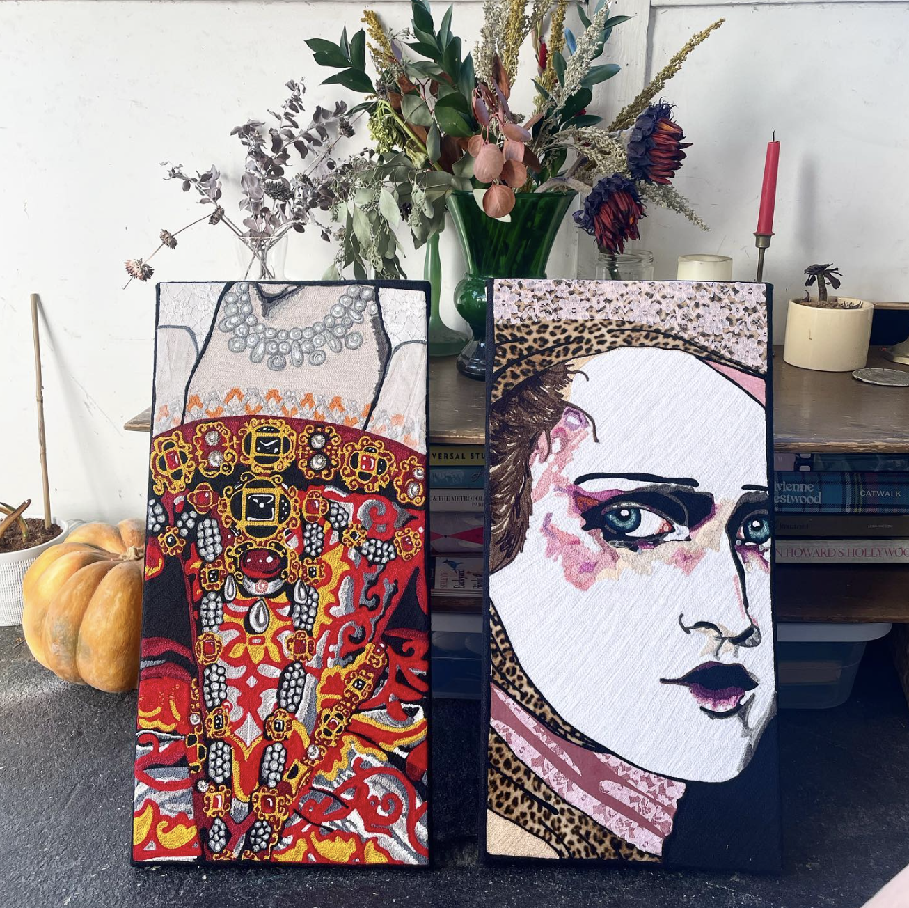 Two embroidered art portraits leaning against a table, with a arrangement of flowers in a green vase and other plants in the background. The left portrait depicts a detailed necklace and intricate ornamental design with red, yellow, black, and white 