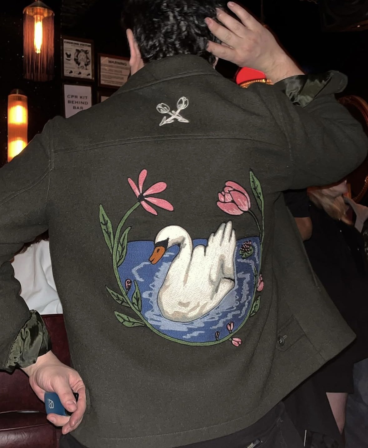 Back of a person's green jacket with a large embroidered design of a swan in a pond, surrounded by pink flowers and green leaves.