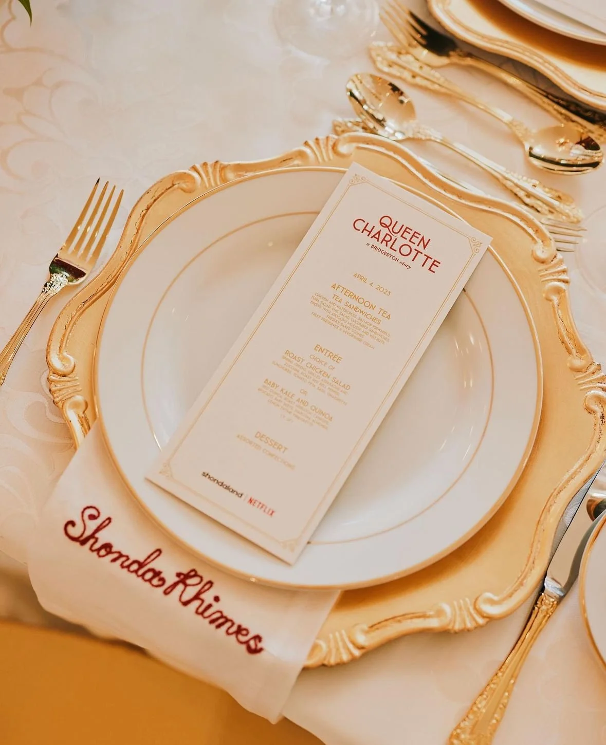 A fancy table setting with a decorative gold-rimmed plate, a fork on the left, and a spoon on the right. A menu card for Queen Charlotte dinner event is placed on the plate, with a cloth napkin underneath reading "Shondos Rhimes."