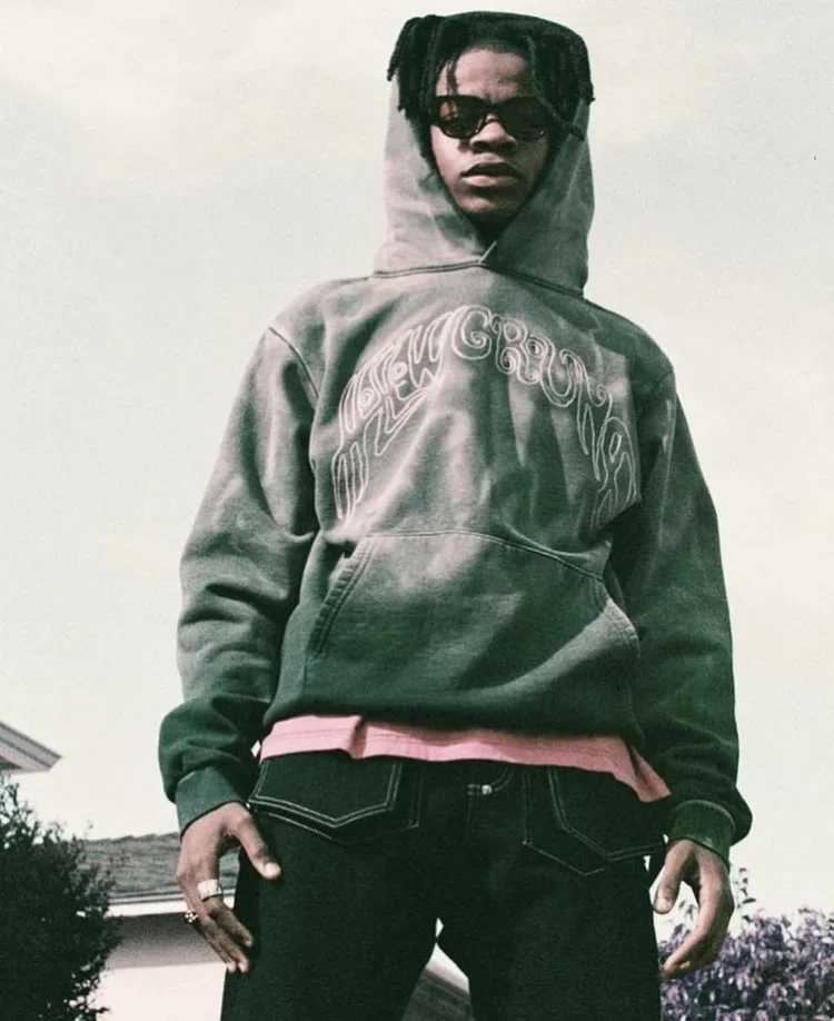 A young individual wearing a gray hoodie with white text, pink shirt underneath, sunglasses, and black jeans, standing outdoors against a plain sky background.