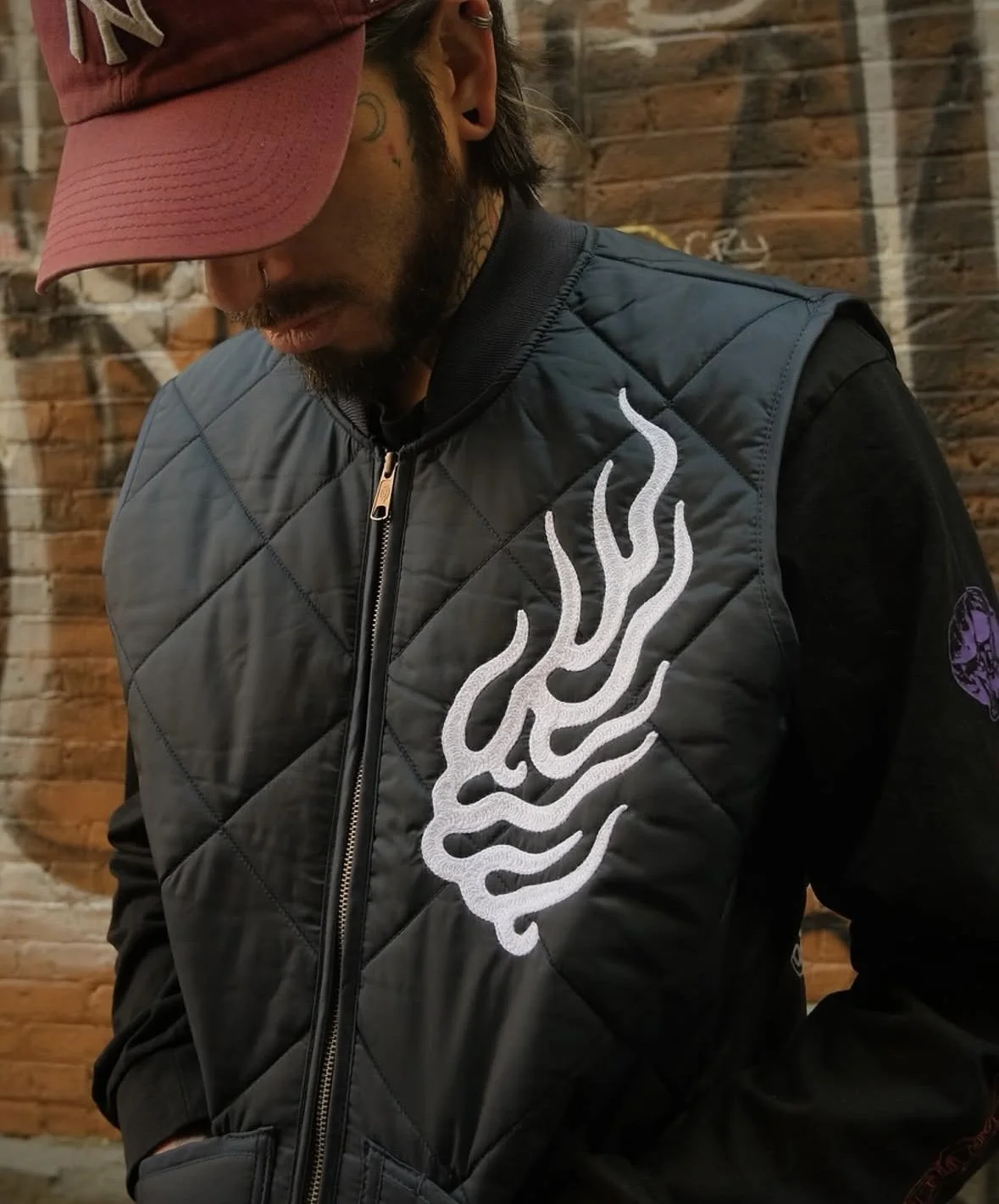 A man wearing a pink and black cap, black jacket with a white flame design on the front, standing against a brick wall.