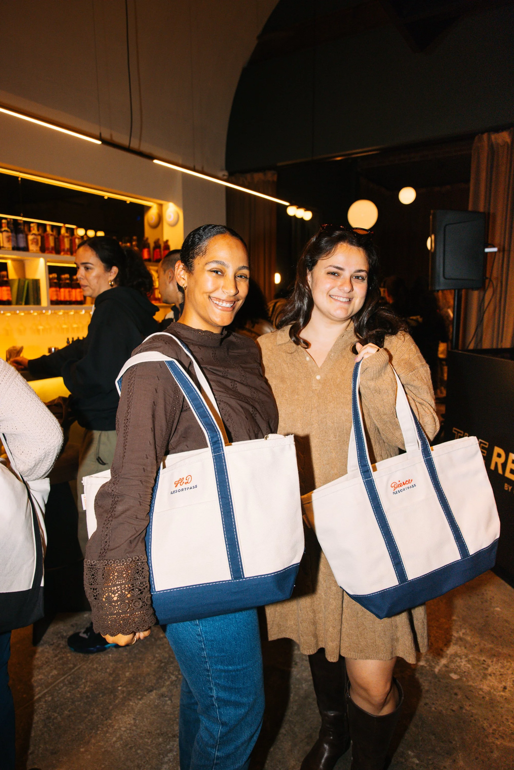 Two women smiling while holding large white and navy tote bags at an indoor event, with bars and other people in the background.