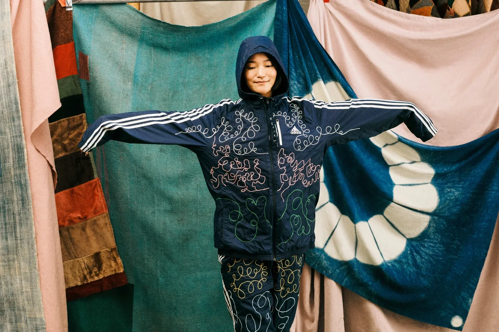 Person wearing a blue Adidas tracksuit standing with arms outstretched in front of colorful fabric curtains. The tracksuit is chainstich embroidered with swirly lines.