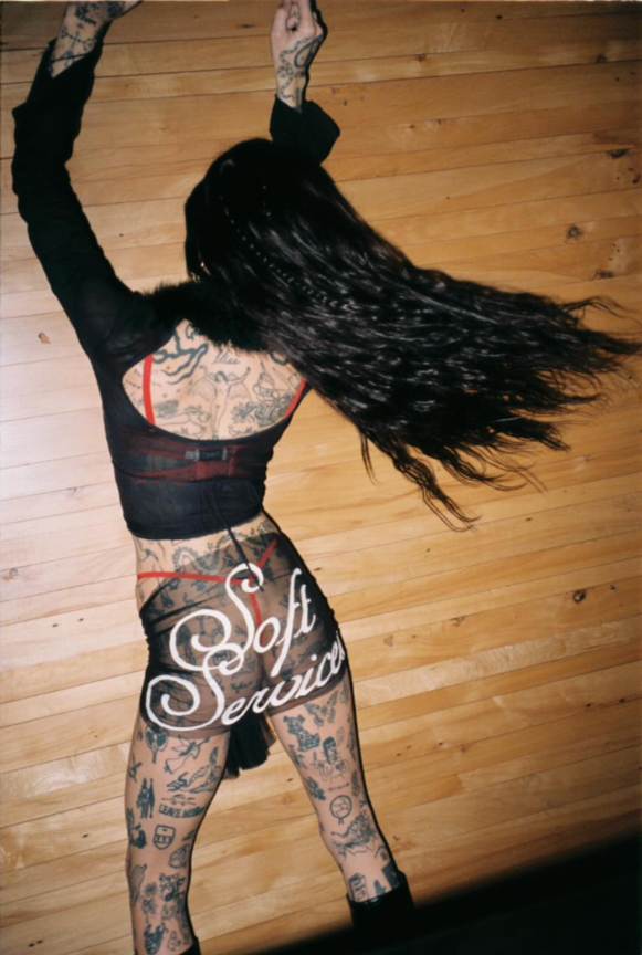A woman with long black hair, tattoos on her back and arms, wearing a black and red mesh lingerie set, stands against a wooden wall with her arms raised.