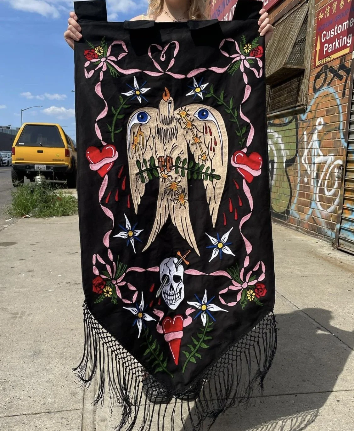 A person holding a black chainstitched embroidered banner with a tattoo-inspired design featuring a bleeding angel wing with a candle wick, surrounded by hearts, skull, flowers, and stars, with pink fringe at the bottom.