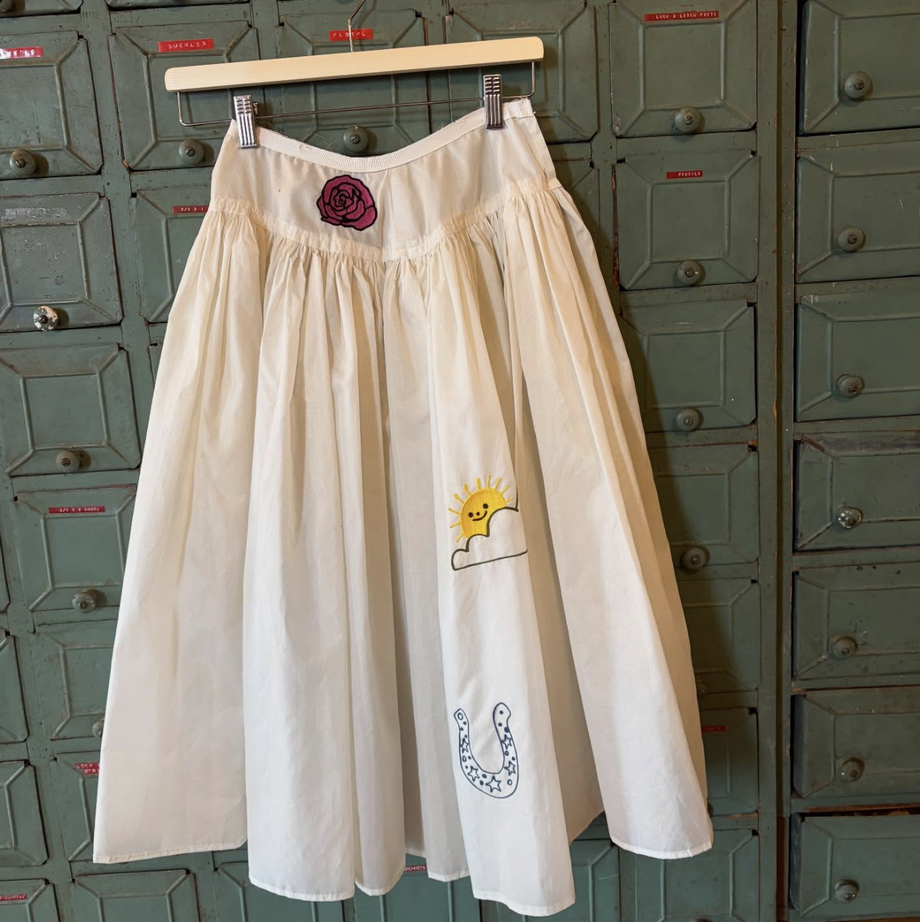 Long cream-colored skirt with whimsical embroidery of a smiling sun and cloud, a giraffe, a pink rose patch near the waistband, displayed on a hanger against a green locker background.