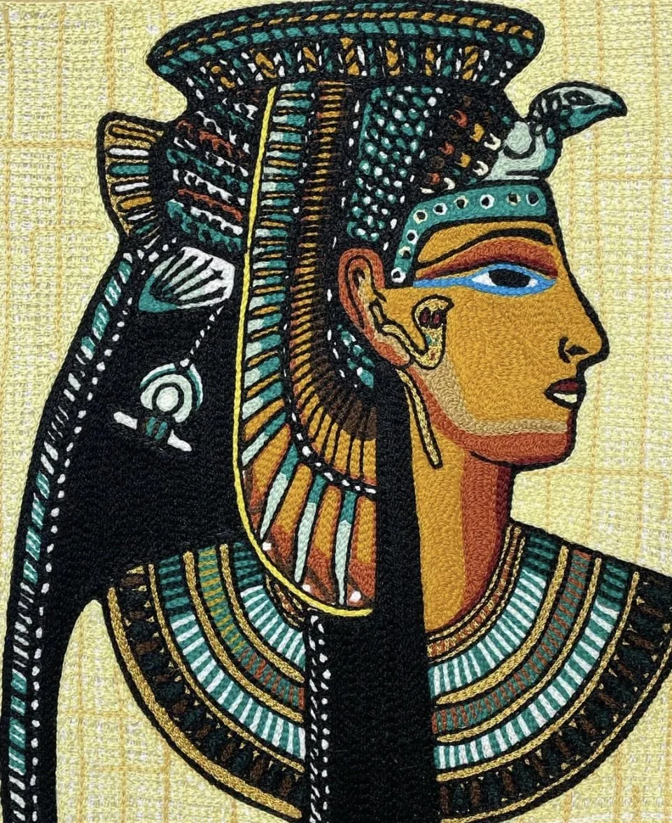 An Egyptian-style embroidered textile depicting a profile of a woman wearing a headdress and jewelry with vibrant colors and intricate patterns.