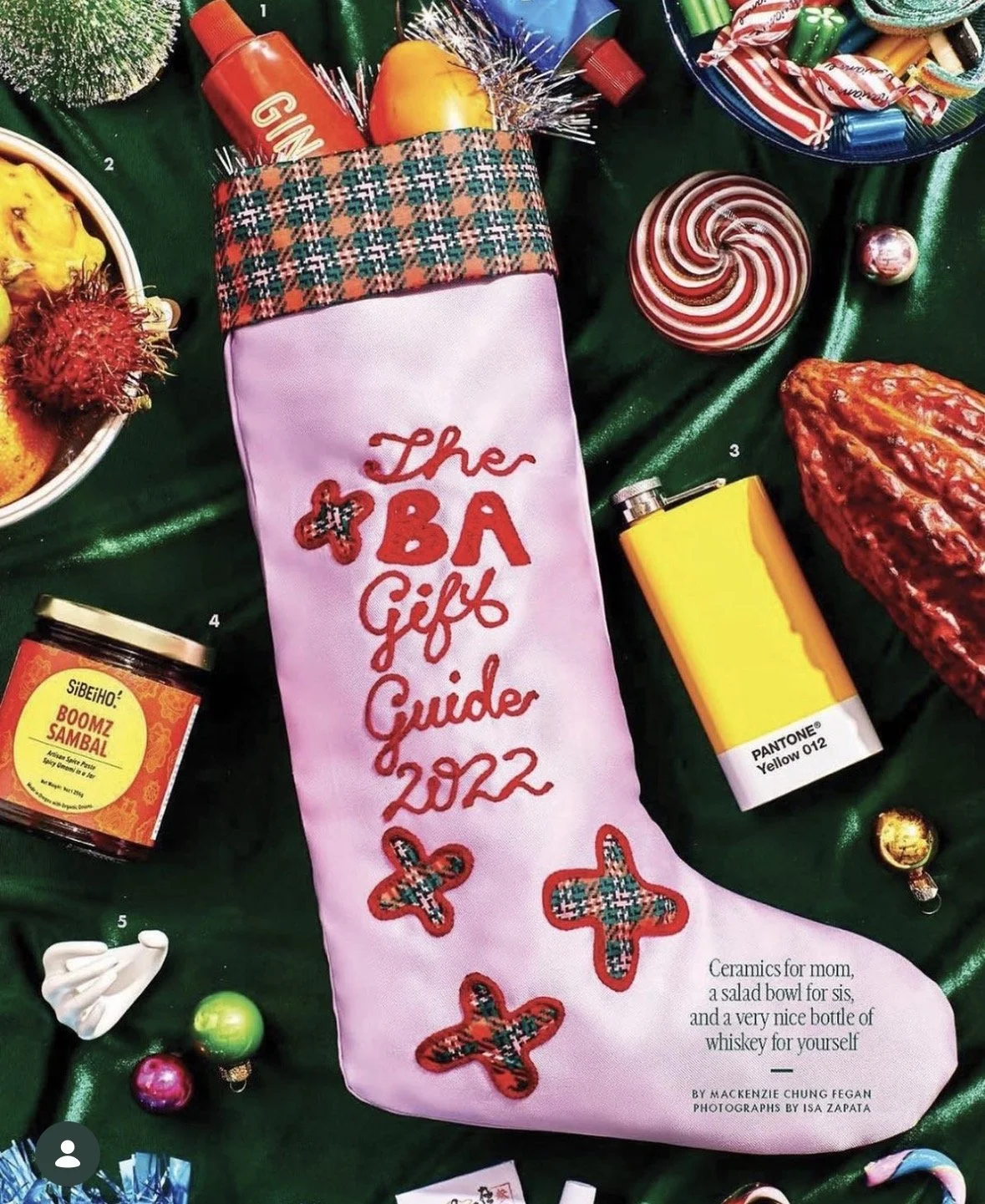 A holiday-themed gift guide booklet titled 'The ABA Gifts Guide 2022' with decorative patches, surrounded by Christmas candies, a yellow perfume bottle, a red and white striped ornament, a small bowl of candies, a jar of spicy jam, and a large dried red chili pepper, all arranged on a green satin cloth.