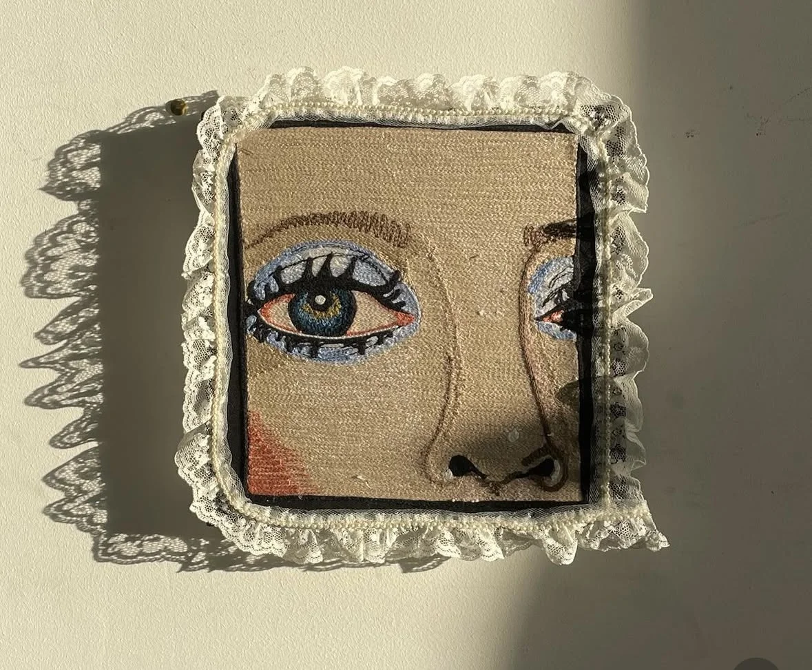 Embroidery artwork of a stylized human face with one eye open and one eye closed, framed with lace, casting a shadow on a plain wall.