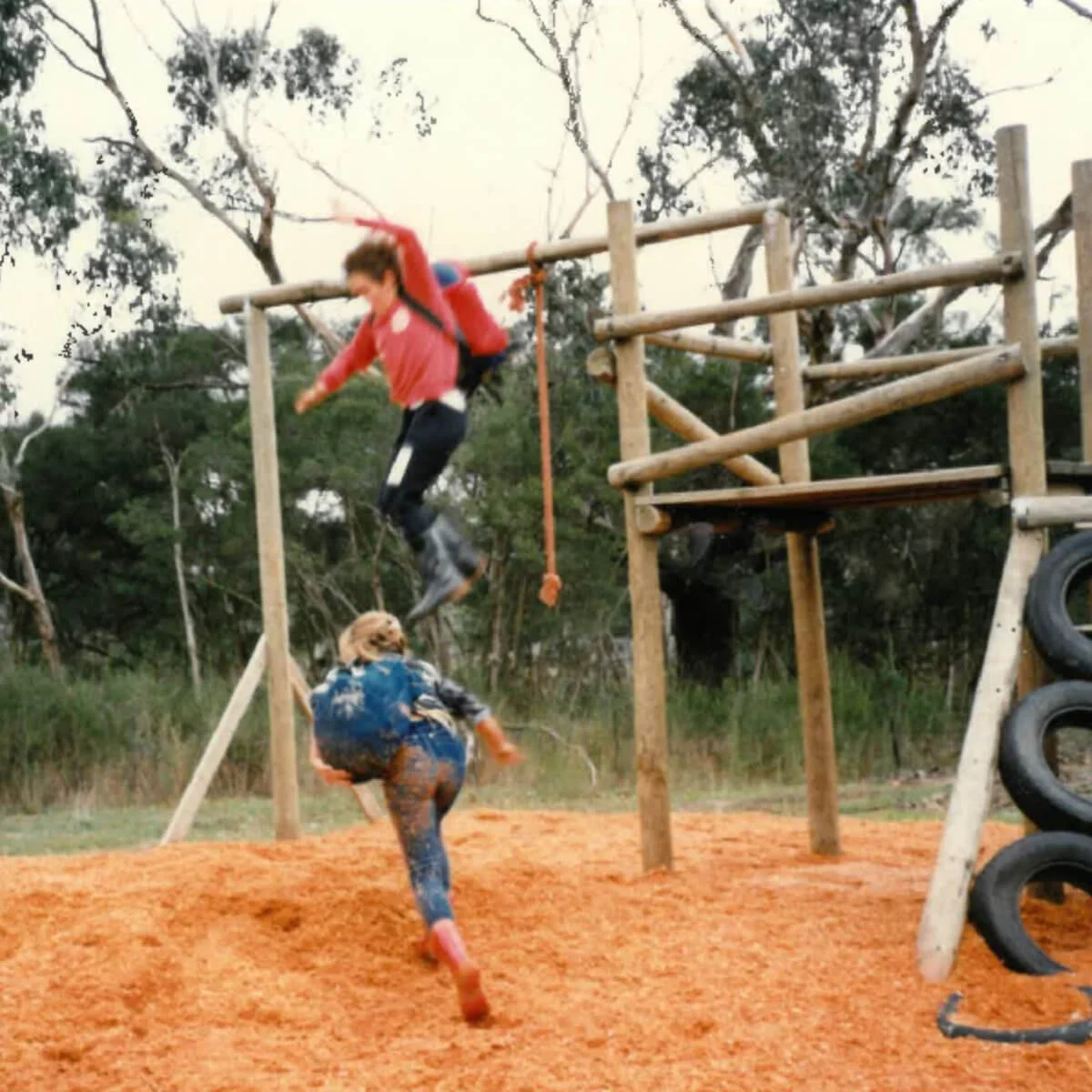 Two people navigating an outdoor obstacle course made of wooden structures and tires, with one person jumping and another climbing. The ground is covered in woodchips, and trees are visible in the background.
