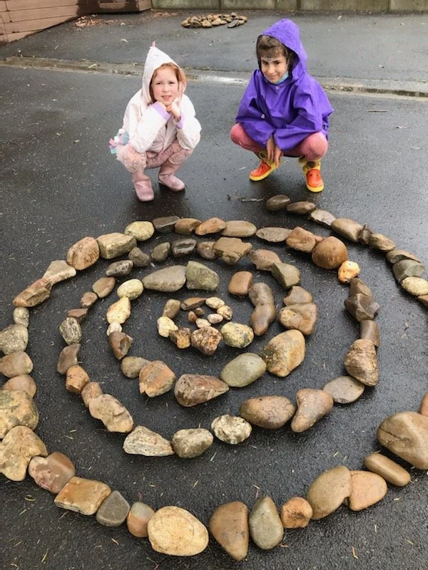 Two children in raincoats squatting next to a spiral pattern made of stones on wet pavement.
