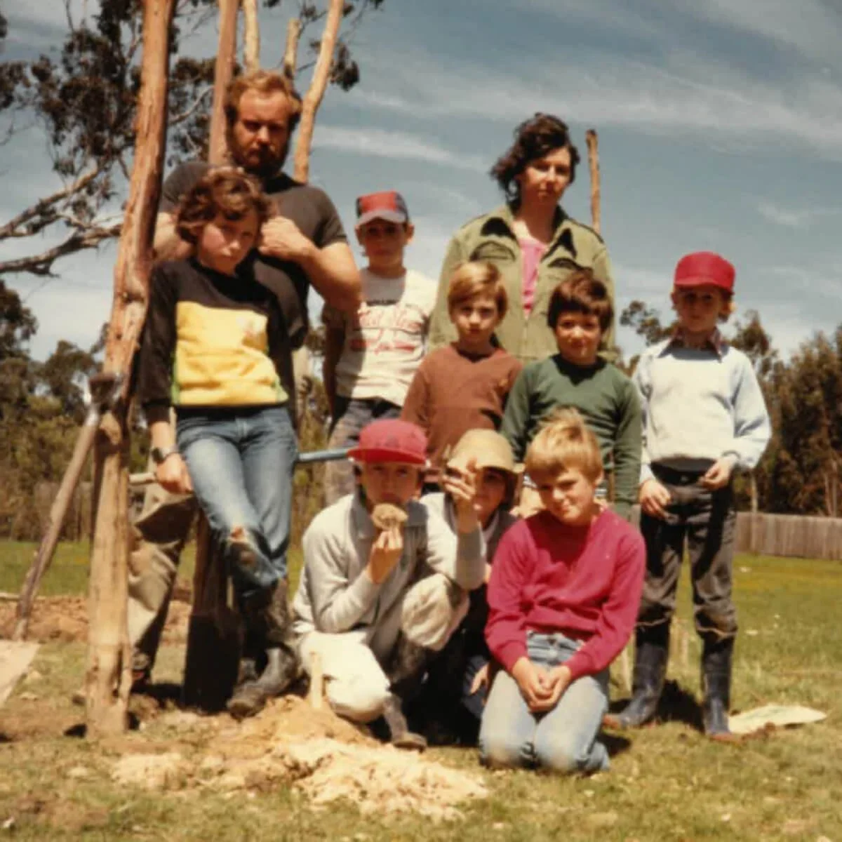 Group of children and two adults outdoors on a grassy field with trees in the background, standing near wooden poles.